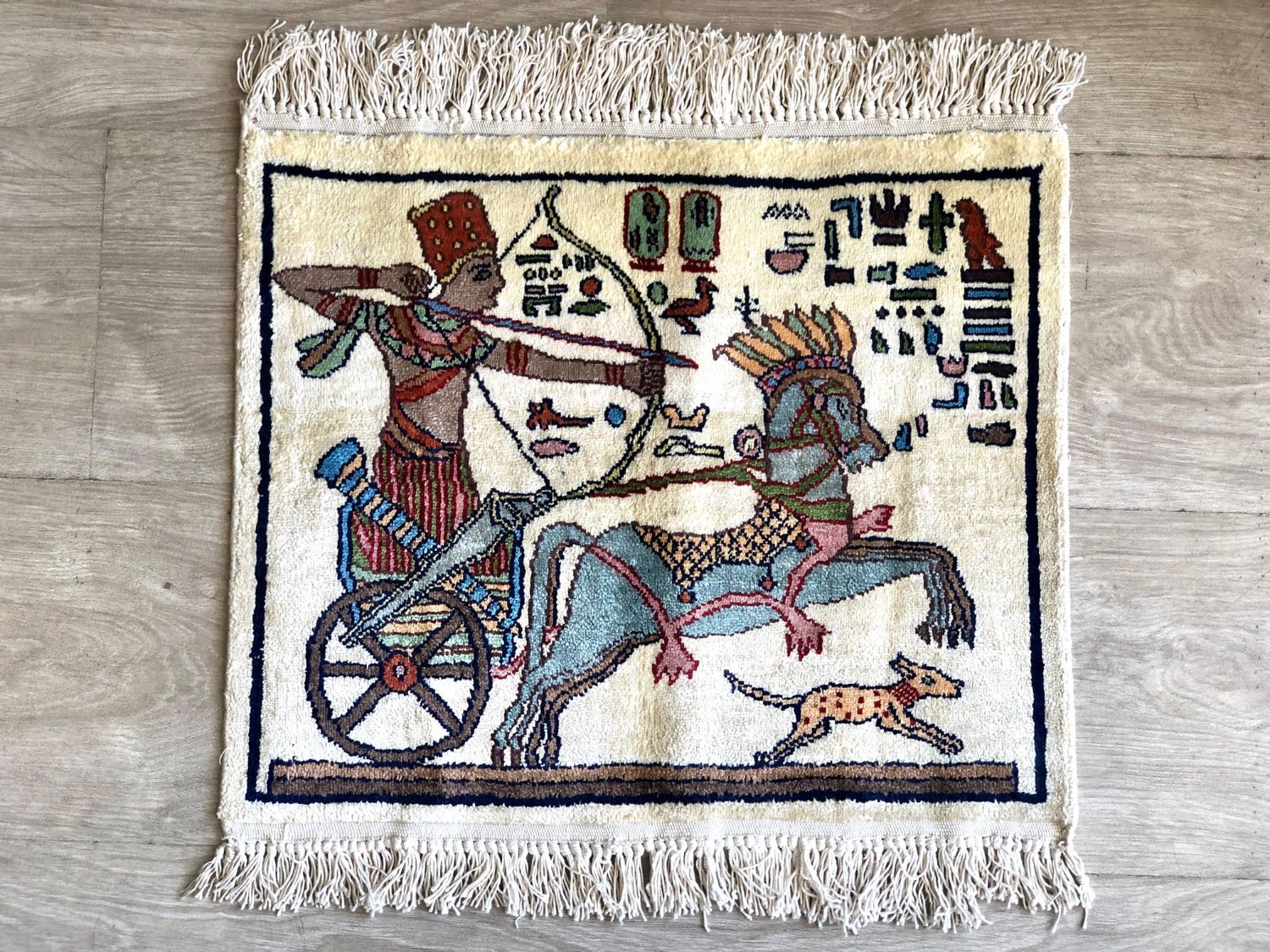 سجادة صغيرة تصويرية تُظهر رامي سهام على عربة يجرها حصان ومعه كلب يركض ورموز على طراز الهيروغليفية، على أرضية كريمية